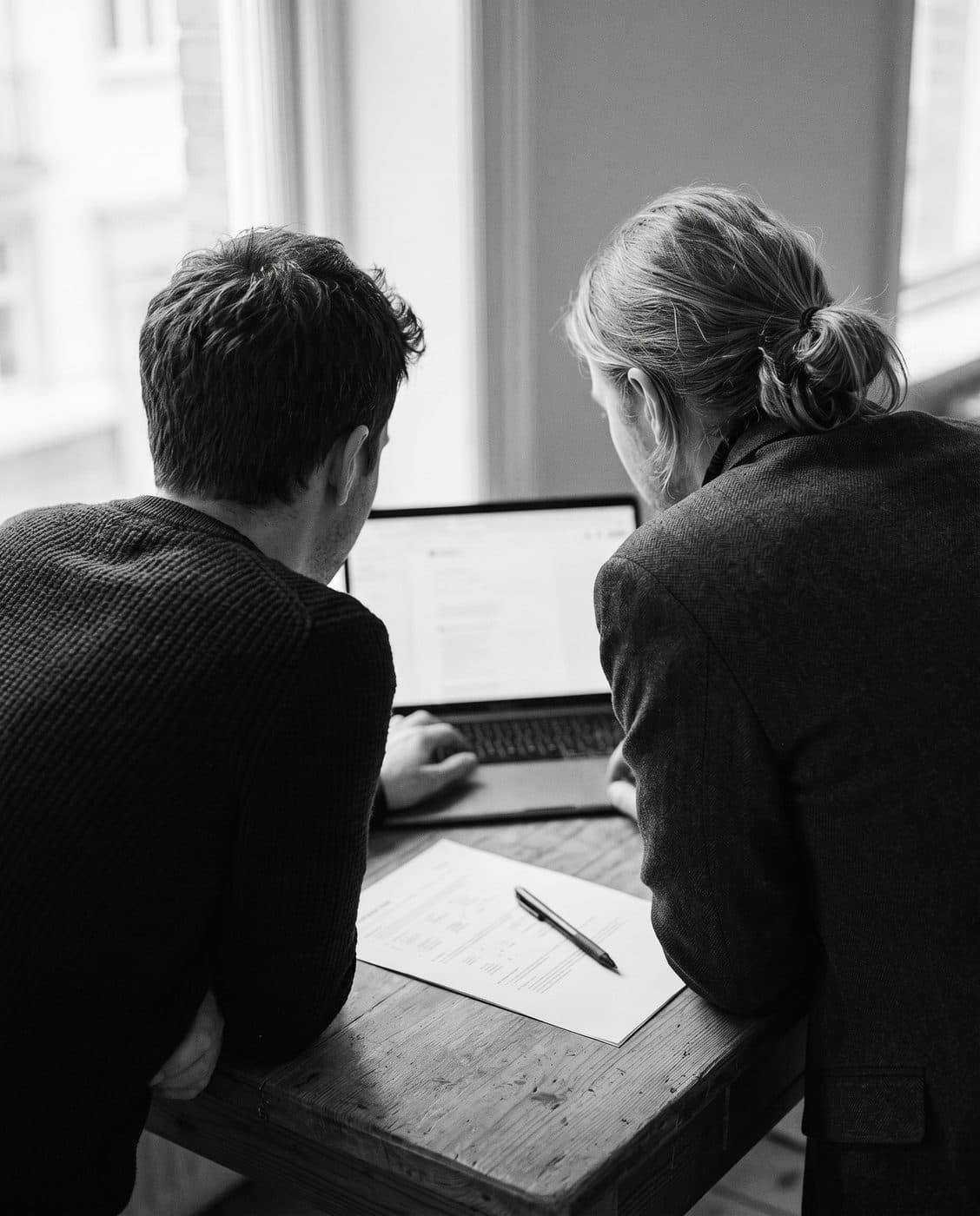 Two colleagues seen from behind, side by side at a table, reading the same printed report, black and white