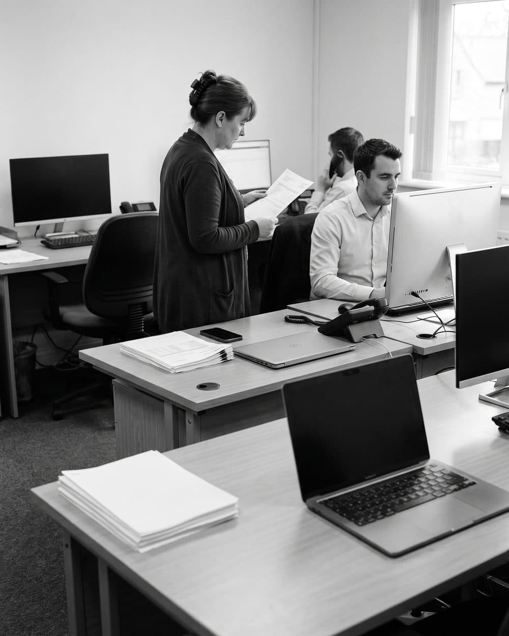 A modest financial operations office, two short rows of desks receding toward a tall window, one person reading a printed statement, black and white