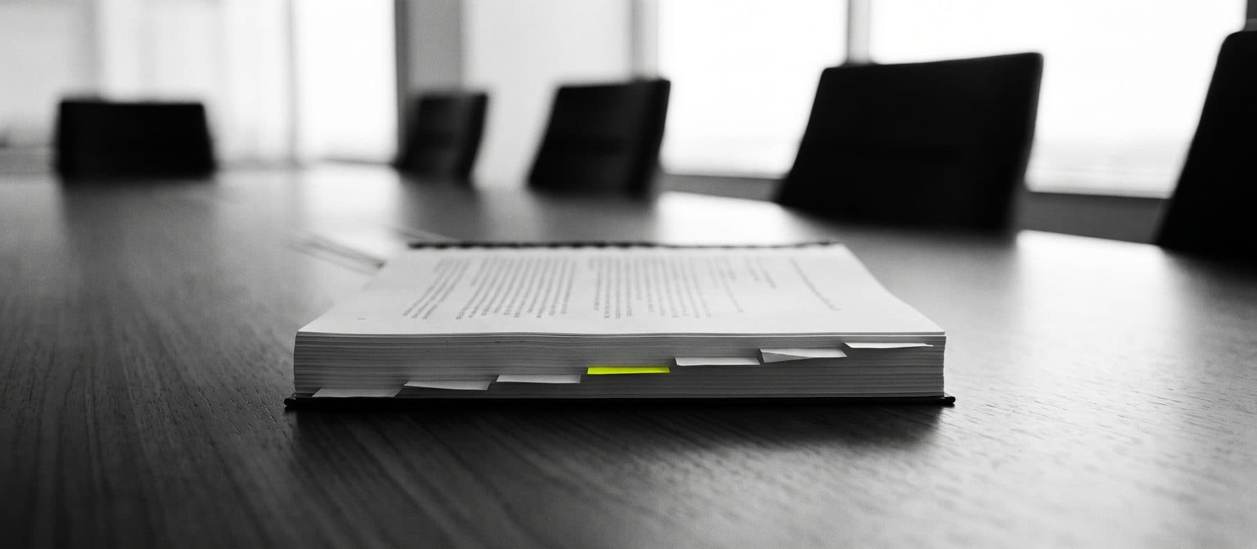 A bound review document alone on a long boardroom table, page markers along its fore edge and one marker in yellow, empty chairs beyond, black and white
