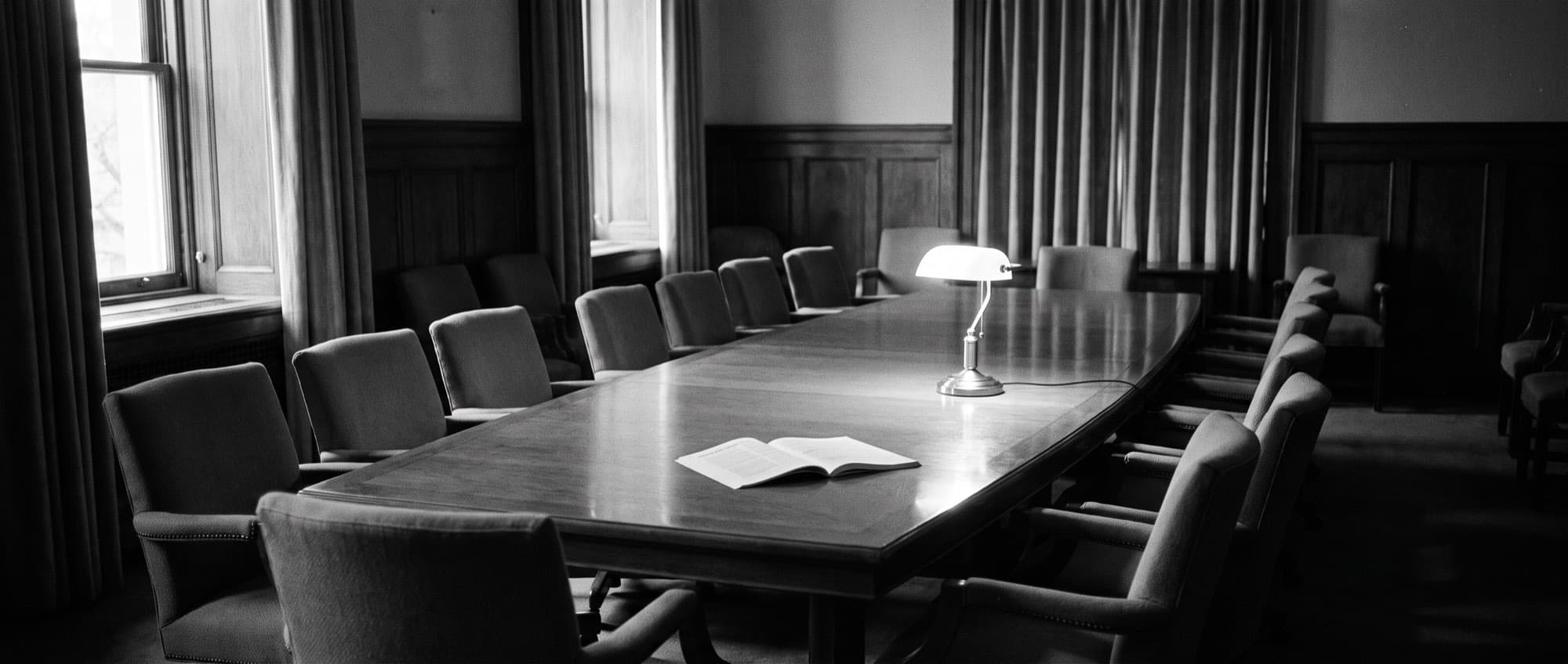An empty boardroom after hours, one printed report open on the table under a desk lamp, black and white